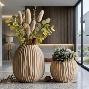 May include: Two beige, textured vases with organic shapes. One vase holds tall, dried grasses and green foliage. The other vase contains a variety of succulents. The vases are on a speckled gray floor.