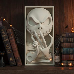 May include: A white relief sculpture of Jack Skellington from The Nightmare Before Christmas. The sculpture is in a rectangular frame and features Jack's face and skeletal hand. Halloween decor with books, a black cat, and string lights.