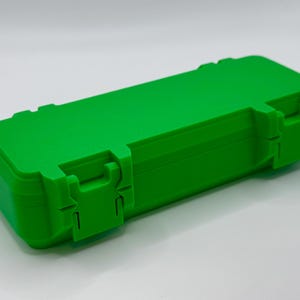 K&ouml;nnte beinhalten: Eine leuchtend gr&uuml;ne, rechteckige Box mit einem Klappdeckel und zwei sicheren Verschl&uuml;ssen. Die Box besteht aus einem soliden, haltbaren Material und ist zum Schutz des Inhalts konzipiert. Die Box wird vor einem wei&szlig;en Hintergrund gezeigt.