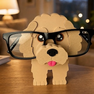 Peut inclure: Un porte-lunettes en forme de chien de couleur beige, fabriqué en matériau superposé. La figurine présente un nez noir, une petite langue rose et de grands yeux sombres. Une paire de lunettes à monture noire repose sur le museau du chien.