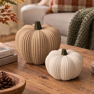 May include: Two decorative pumpkins with a ribbed, textured surface. The larger pumpkin is beige, and the smaller one is off-white. Both feature a dark green, faceted stem. The pumpkins are positioned on a wooden surface, showcasing a rounded shape.