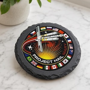 May include: A round, dark gray slate coaster featuring a circular graphic with the text PROJECT HAIL MARY. The design includes a central white spacecraft illustration, a bright yellow sunburst, and a border of various international flags.