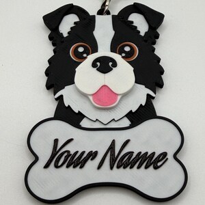 Può includere: Un portachiavi a forma di cane bianco e nero con un'etichetta a forma di osso. Il cane ha occhi marroni, una lingua rosa e il testo "Your Name" in corsivo sull'etichetta dell'osso. È attaccato un portachiavi argentato.