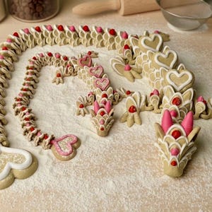 May include: Dragon-shaped cookies decorated with pink, red, and white icing. The cookies are arranged on a lightly dusted surface, with heart-shaped accents and a whimsical design. A creative and festive treat.