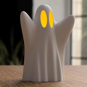 Może przedstawiać: Biała lampa dekoracyjna w kształcie ducha z dwoma żółtymi owalnymi światłami jako oczami. Lampa ma drapowany design i stoi na drewnianej powierzchni. Tło jest rozmyte.