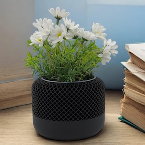 May include: A black speaker with a mesh design and a white flower arrangement in the top. The speaker is sitting on a wooden surface.