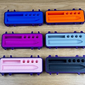Può includere: Sei custodie rettangolari colorate con cornici viola. Ogni custodia contiene uno stilo e scomparti per accessori. I colori includono blu, arancione, viola, azzurro, rosa e nero. Le custodie sono disposte su una superficie di legno.