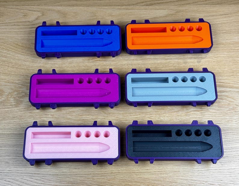 K&ouml;nnte beinhalten: Sechs farbenfrohe Stifteetuis in Blau, Orange, Lila, Hellblau, Rosa und Schwarz. Jedes Etui hat einen lila Rahmen und enth&auml;lt einen stiftf&ouml;rmigen Ausschnitt und mehrere kleine kreisf&ouml;rmige Ausschnitte. Die Etuis liegen auf einer Holzoberfl&auml;che.