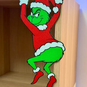 May include: A green cartoon character in a red Santa suit and hat with white trim, hanging from a wooden shelf. The character has a mischievous grin and is wearing red boots. A festive Christmas decoration.