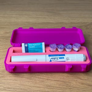 May include: A magenta case, open to reveal a white and blue Wegovy FlexTouch pen, pre-injection swabs, and small purple containers. The case is on a wooden surface.