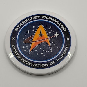 May include: A white, circular Starfleet Command badge with a blue and orange emblem. The emblem features a stylized star and the text "STARFLEET COMMAND" and "UNITED FEDERATION OF PLANETS".