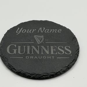 May include: A round, dark gray slate coaster with the Guinness logo and the text "Your Name" engraved on it. The coaster also features the words "Guinness Draught" and the year "1759".