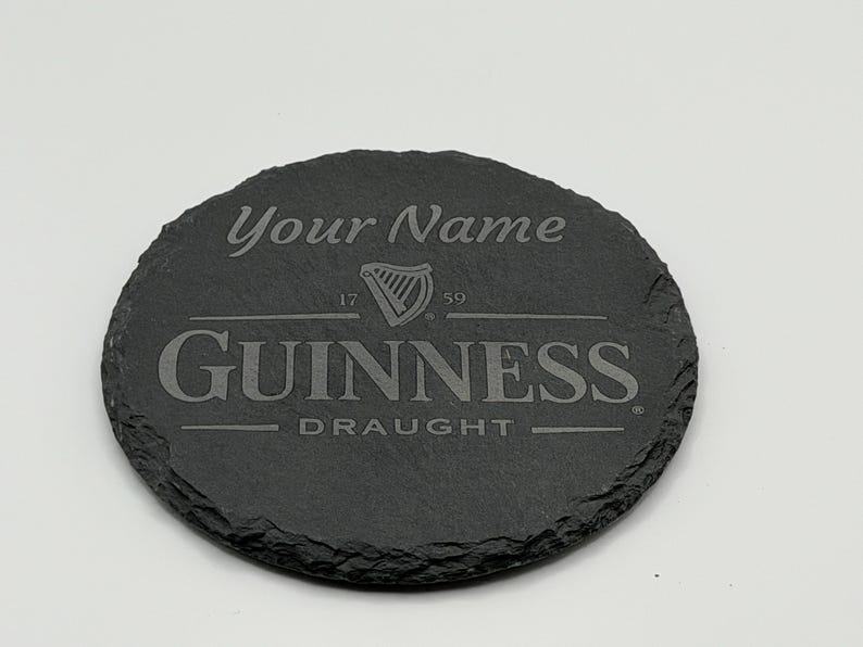 May include: A round, dark gray slate coaster with the Guinness logo and the text "Your Name" engraved on it. The coaster also features the words "Guinness Draught" and the year "1759".