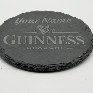 May include: Oval, dark gray slate coaster with the Guinness logo and the words "Your Name" engraved at the top. The Guinness logo includes a harp and the words "GUINNESS DRAUGHT".
