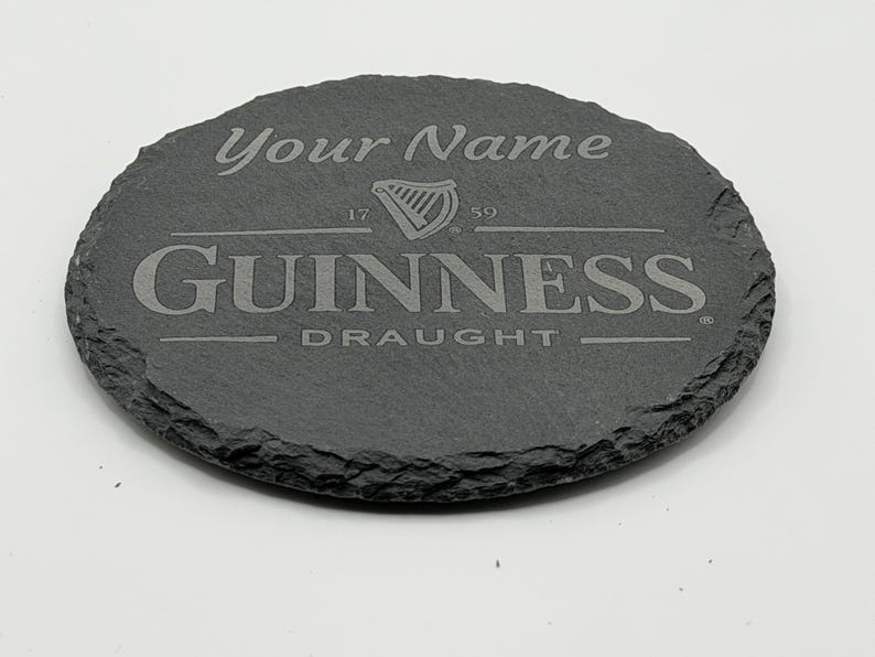 May include: Oval, dark gray slate coaster with the Guinness logo and the words "Your Name" engraved at the top. The Guinness logo includes a harp and the words "GUINNESS DRAUGHT".
