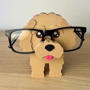 May include: A tan-colored dog-shaped eyeglasses holder with black-framed glasses. The dog has a black nose, pink tongue, and brown eyes. The holder is made of a light brown material and is designed to hold eyeglasses.