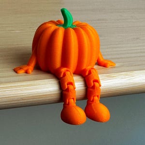 May include: A 3D-printed orange pumpkin ornament with articulated legs and arms, and a green stem. The pumpkin is sitting on a wooden surface. The figurine is a whimsical decorative item.