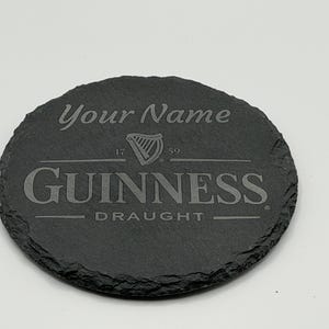 May include: A round, dark gray slate coaster with the Guinness logo and the text "Your Name". The coaster features the Guinness harp logo and the words "Guinness Draught". The coaster is approximately 4 inches in diameter.