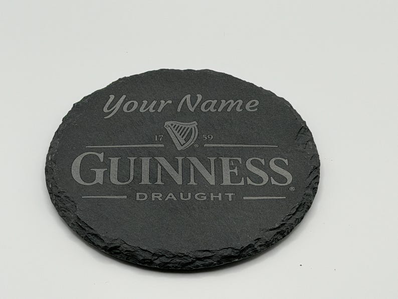 May include: A round, dark gray slate coaster with the Guinness logo and the text "Your Name". The coaster features the Guinness harp logo and the words "Guinness Draught". The coaster is approximately 4 inches in diameter.