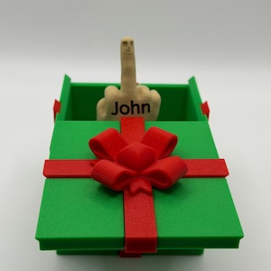 May include: A green gift box with a red ribbon and bow, containing a beige figurine of a hand making a middle finger gesture. The hand features a small face on the fingertip and the name John printed in black letters on the base of the hand.