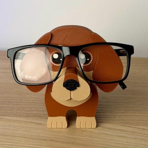 Może przedstawiać: Brązowo-beżowy uchwyt na okulary w kształcie jamnika z czarnymi oprawkami. Uchwyt ma tr&oacute;jwymiarowy design z beżowym pyskiem i łapami oraz brązowymi uszami i korpusem. Okulary spoczywają na nosie psa.