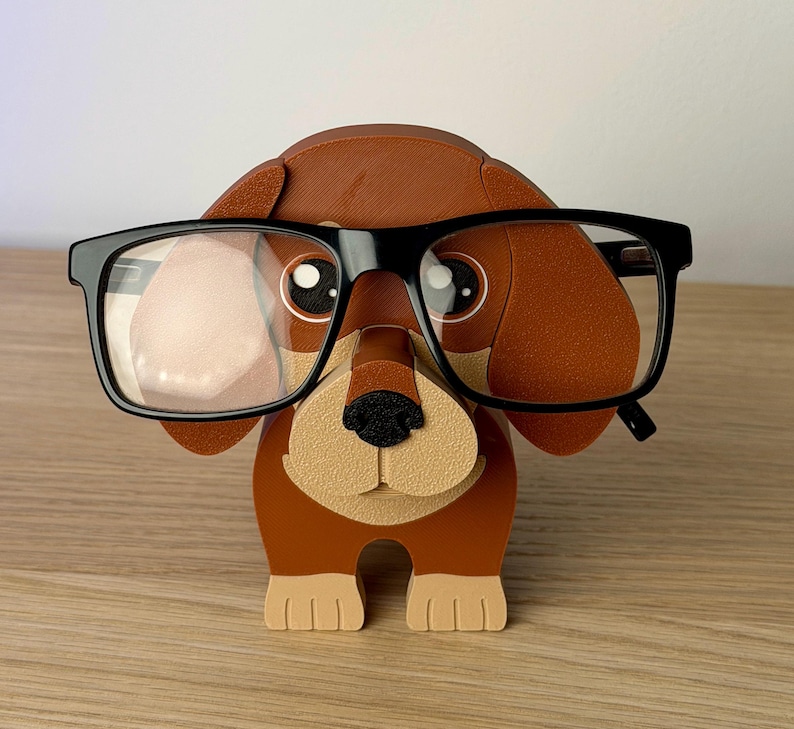 Może przedstawiać: Brązowo-beżowy uchwyt na okulary w kształcie jamnika z czarnymi oprawkami. Uchwyt ma tr&oacute;jwymiarowy design z beżowym pyskiem i łapami oraz brązowymi uszami i korpusem. Okulary spoczywają na nosie psa.
