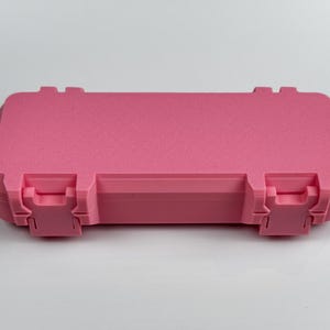 K&ouml;nnte beinhalten: Ein rosa rechteckiger Koffer mit strukturierter Oberfl&auml;che. Der Koffer hat zwei Verschl&uuml;sse vorne und zwei hinten. Der Koffer ist geschlossen und liegt auf einer wei&szlig;en Oberfl&auml;che.