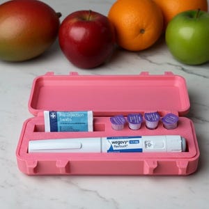 Puede incluir: Un estuche rosa abierto que contiene un bolígrafo de inyección Wegovy FlexTouch, toallitas previas a la inyección y pequeños recipientes morados. Frutas, como un mango, una manzana, una naranja y una manzana verde, en el fondo.