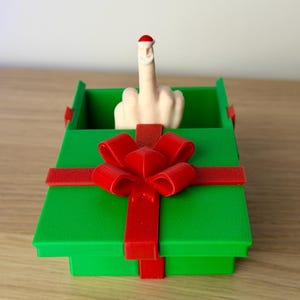Könnte beinhalten: Eine grüne Geschenkbox mit roter Schleife und Band, gekrönt von einer Hand, die den Mittelfinger zeigt. Der Finger trägt einen Weihnachtsmannhut. Die Box steht auf einer Holzoberfläche.