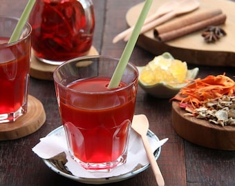 May include: Two glasses of red liquid with straws, a wooden chopping board with cinnamon sticks, star anise, and sugar cubes, and a bowl of dried ginger and turmeric.