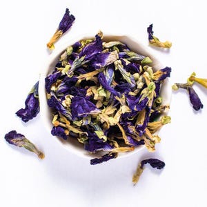 May include: A white bowl containing dried butterfly pea flowers, displaying a striking blue colour, with petals scattered around. The flowers have blue and yellow tones, set against a bright white background.