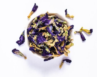 May include: A white bowl containing dried butterfly pea flowers, displaying a striking blue colour, with petals scattered around. The flowers have blue and yellow tones, set against a bright white background.