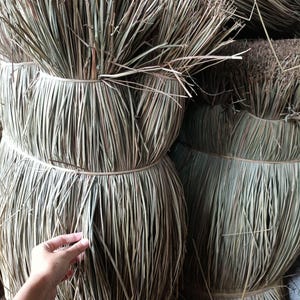 Puede incluir: Grandes manojos de tallos de hierba natural seca de color marrón claro, atados con hilo fino. Las hojas largas, finas y fibrosas están dispuestas verticalmente en forma cilíndrica, creando una apariencia densa y texturizada con tonos tierra.