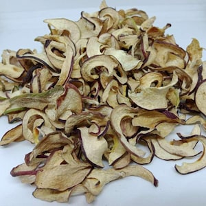 Könnte beinhalten: Nahaufnahme von getrockneten Apfelscheiben, die eine Mischung aus Beige, Braun und Rot-Violett aufweisen. Die dehydrierten Fruchtstücke haben gewellte Ränder und unterschiedliche Größen, was auf ein natürliches, hausgemachtes Produkt hindeutet. Das Bild hebt die Textur und die Farbvariationen hervor.