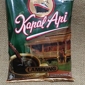 Puede incluir: Un paquete de café verde y dorado con las palabras "Kapal Api" en escritura blanca. El paquete presenta una ilustración de un barco y una taza de café. La palabra "LAMPUNG" también es visible.