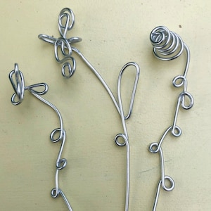 May include: Several handmade silver wire flower sculptures with various designs. The flowers have looped and swirled details, with stems of varying lengths. The sculptures are displayed on a light yellow surface.