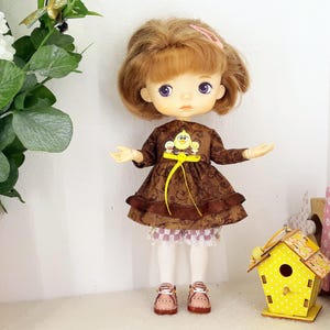 May include: A brown and white doll with blonde hair wearing a brown dress with a yellow bird design and a yellow ribbon. The doll is wearing white tights and brown shoes. The doll is standing on a white surface in front of a yellow birdhouse.