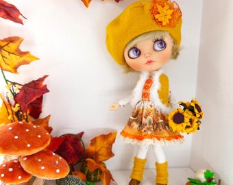 May include: A doll with blonde hair and purple eyes wearing a mustard yellow beret with an orange leaf accent, a white faux fur vest, and a dress featuring an autumn-themed print. The doll wears white leggings, yellow leg warmers, and brown shoes.