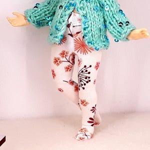 May include: White tights with a floral pattern in shades of orange and brown. The tights are worn by a doll with a teal knitted cardigan.