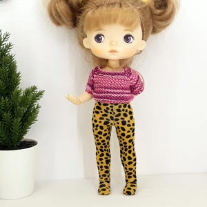May include: A doll wearing a pink and white striped sweater and yellow and black leopard print leggings.