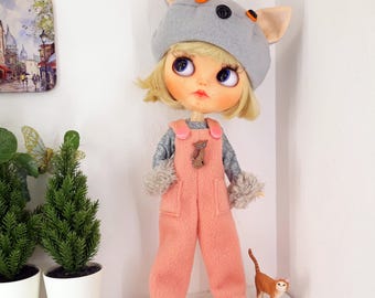 May include: A doll dressed in a grey cat-eared hat, a grey long-sleeved top, and peach-coloured dungarees. The doll has blonde hair and stands beside a small orange cat figurine. The outfit includes fluffy cuffs and a small bear-shaped button.