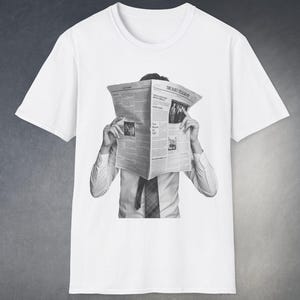 May include: White t-shirt with a black and white graphic of a person with a newspaper obscuring their face. The newspaper displays the text "THE DAILY TELEGRAPH". The person is wearing a white shirt and tie, creating a monochrome design.