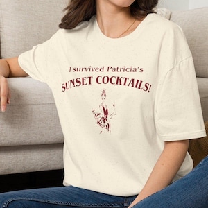 May include: A cream-colored short-sleeve t-shirt with a speckled texture. The front features dark red text that reads I survived Patricia's SUNSET COCKTAILS! above a stylized dark red graphic illustration of a person holding a drink.