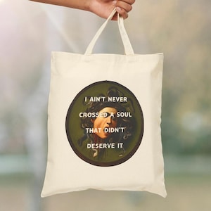 May include: A cream-colored tote bag featuring a circular graphic of Medusa with the text "I ain't never crossed a soul that didn't deserve it." The bag has two cream-colored handles.