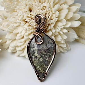 May include: A copper wire wrapped pendant featuring a green, brown, and white moss agate stone. The pendant is shaped like a teardrop and has a copper wire bail.