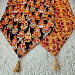 May include: A reversible table runner with a black background featuring a pattern of gnomes with pumpkins and fall leaves. The other side of the runner is a gold and orange pattern. The runner has tassels on each end.