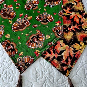 May include: A green and brown table runner with a Thanksgiving theme. One side features a pattern of turkeys playing football with footballs and leaves. The other side features a pattern of brown, orange, and yellow leaves. The runner has brown tassels on each end.