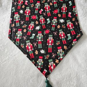 May include: Black Christmas table runner with a repeating pattern of toy soldiers in red, green, and white. The runner features gingerbread men, stockings, and poinsettias. A green tassel hangs from the bottom.