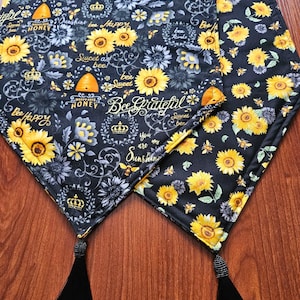 May include: Black table runner with a sunflower and bee pattern. The runner features yellow sunflowers, gray flowers, bees, and text that reads "Bee Grateful" and "Sweet as Bee". Black tassels adorn the ends.