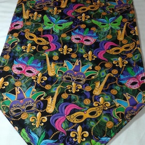 May include: A black table runner with a Mardi Gras theme. The runner features a repeating pattern of gold, purple, green, and pink masks, fleur-de-lis, and musical notes. The runner has a tassel at the end.